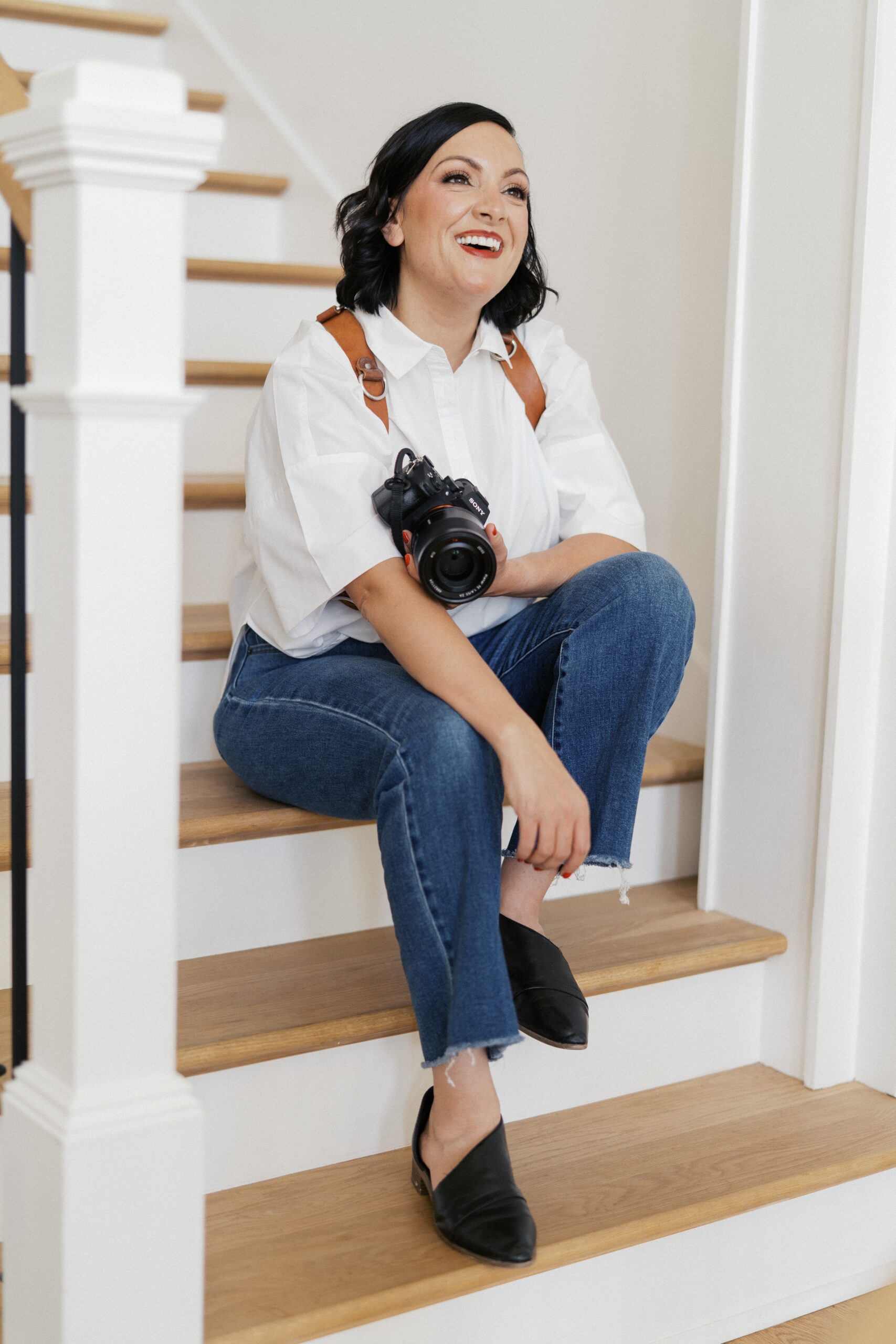 a nashville family photographer is sitting on a stairwell smiling while holding her camera 